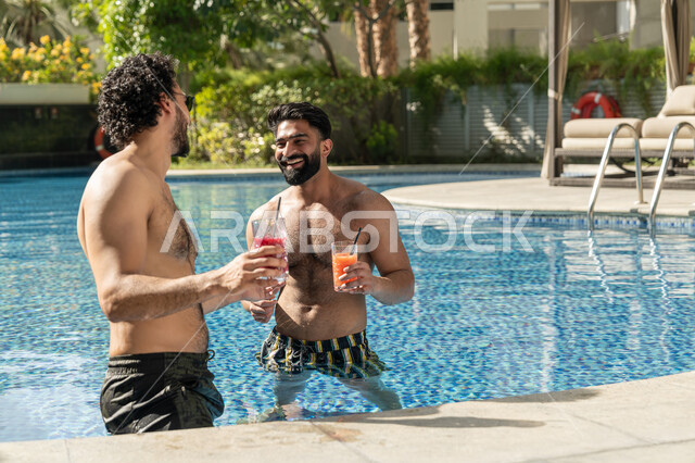قضاء العطلة الصيفية في المنتجعات السياحية, مشاركة الأصدقاء اللحظات الممتعة, القيام بالأنشطة الترفيهية المسلية, شابان عربيان خليجيان إماراتيان يجلسان داخل حوض السباحة في إحدى الفنادق بالإمارات, الاستمتاع بالمشروبات الباردة المنعشة