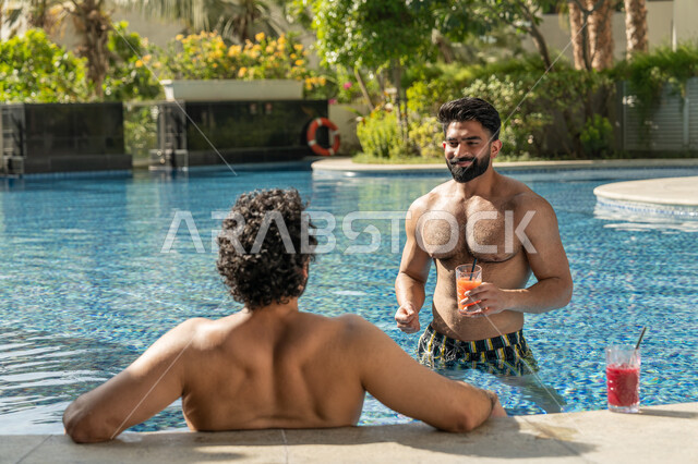 مشاركة الأصدقاء اللحظات الممتعة, قضاء العطلة الصيفية في المنتجعات السياحية, القيام بالأنشطة الترفيهية المسلية, شابان عربيان خليجيان إماراتيان يجلسان داخل حوض السباحة في إحدى الفنادق بالإمارات, الاستمتاع بالمشروبات الباردة المنعشة