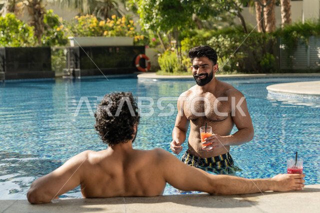 Doing fun recreational activities, sharing enjoyable moments with friends, spending summer vacation in tourist resorts, two young Emirati Gulf Arab men sitting in the swimming pool in one of the hotels in the Emirates, enjoying refreshing cold drinks