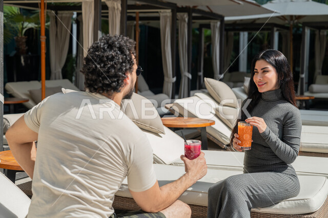 قضاء الأوقات العائلية المسلية, القيام بالأنشطة الترفيهية الممتعة في الإجازة, الاستمتاع بالمشروبات الصيفية الباردة, زوجان عربيان خليجيان إماراتيان يرتديان الزي الكاجوال يجلسان في إحدى المنتجعات في الإمارات