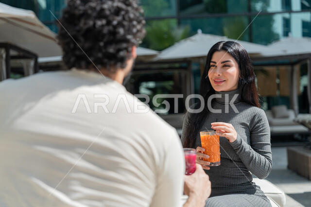 الاستمتاع بالمشروبات الصيفية الباردة, قضاء الأوقات العائلية المسلية, القيام بالأنشطة الترفيهية الممتعة في الإجازة, زوجان عربيان خليجيان إماراتيان يرتديان الزي الكاجوال يجلسان في إحدى المنتجعات في الإمارات