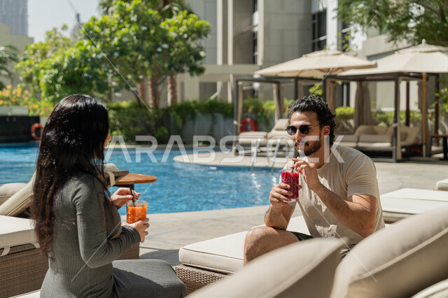 قضاء الأوقات العائلية المسلية, الاستمتاع بالمشروبات الصيفية الباردة, القيام بالأنشطة الترفيهية الممتعة في الإجازة, زوجان عربيان خليجيان إماراتيان يرتديان الزي الكاجوال يجلسان في إحدى المنتجعات في الإمارات