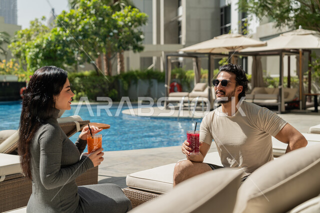 الاستمتاع بالمشروبات الصيفية الباردة, قضاء الأوقات العائلية المسلية, القيام بالأنشطة الترفيهية الممتعة في الإجازة, زوجان عربيان خليجيان إماراتيان يرتديان الزي الكاجوال يجلسان في إحدى المنتجعات في الإمارات