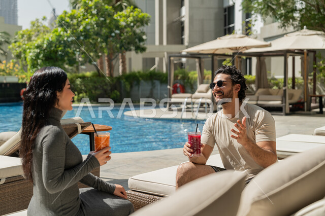 قضاء الأوقات العائلية المسلية, الاستمتاع بالمشروبات الصيفية الباردة, القيام بالأنشطة الترفيهية الممتعة في الإجازة, زوجان عربيان خليجيان إماراتيان يرتديان الزي الكاجوال يجلسان في إحدى المنتجعات في الإمارات