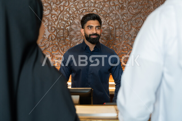 إتمام إجراءات الحجز والدخول, الفنادق والمنتجعات السياحية في الإمارات, قضاء أوقات ممتعة في العطلة الصيفية, صورة مقربة من الخلف لرجل عربي خليجي إماراتي يرتدي الكندورة والغترة البيضاء يقف مع زوجته امام مكتب الاستقبال في الفندق