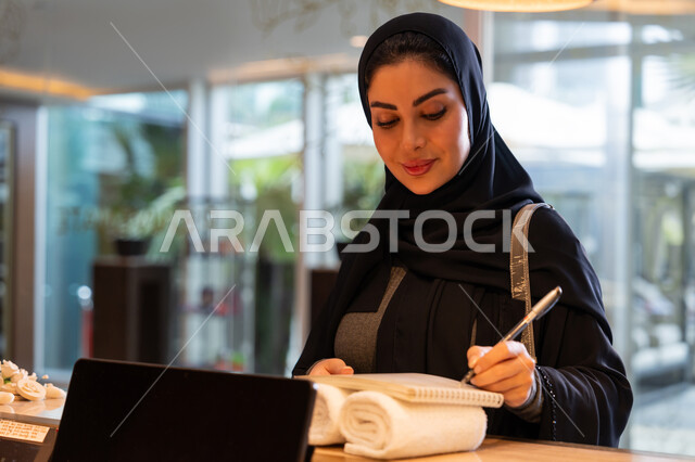 القيام بإجراءات الحجز والدخول, قضاء أوقات ممتعة في العطلة الصيفية, منتجعات وفنادق الإمارات العربية المتحدة, امرأة عربية خليجية إماراتية محجبة ترتدي العباءة السوداء تقوم بتسجيل المعلومات والبيانات الشخصية