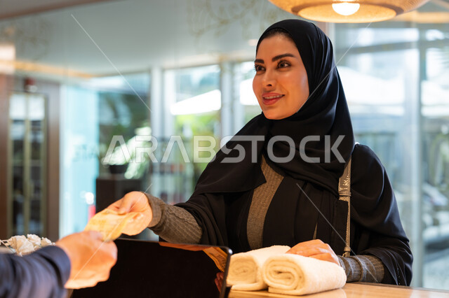 قضاء أوقات ممتعة في العطلة الصيفية, القيام بإجراءات الحجز والدخول, منتجعات وفنادق الإمارات العربية المتحدة, امرأة عربية خليجية إماراتية محجبة ترتدي العباءة السوداء تقوم بالدفع النقدي في مكتب الاستقبال