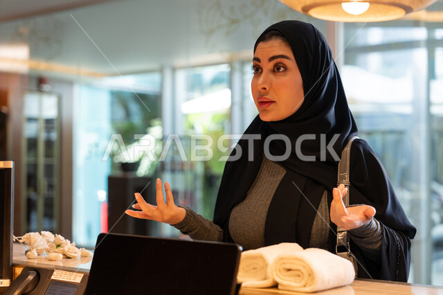 التساؤل والاستفسار عن شيء ما, القيام بإجراءات الحجز والدخول, قضاء العطلة الصيفية في المنتجعات والفنادق, امرأة عربية خليجية إماراتية محجبة ترتدي العباءة السوداء تقف في مكتب الاستقبال وترفع يديها بتعابير الاستفهام