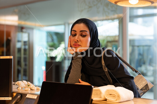 الانتظار وإمهال بعض الوقت, القيام بإجراءات الحجز والدخول, قضاء العطلة الصيفية في المنتجعات والفنادق, امرأة عربية خليجية إماراتية محجبة ترتدي العباءة السوداء تقف في مكتب الاستقبال وتضع يدها على خدها بتعابير الملل