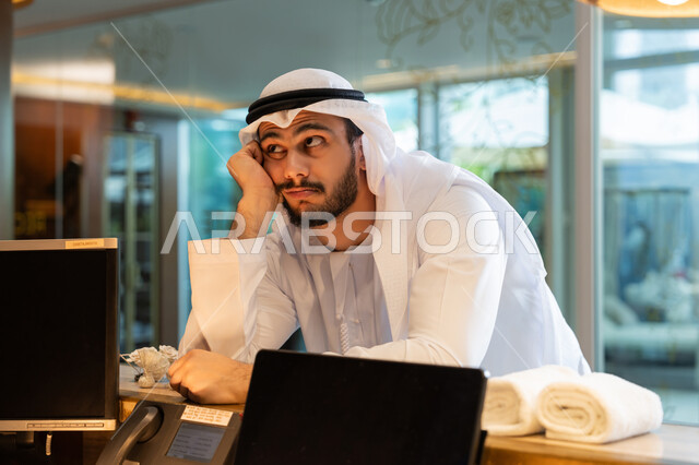 الانتظار وإمهال بعض الوقت, القيام بإجراءات الحجز والدخول, قضاء العطلة الصيفية في المنتجعات والفنادق, رجل عربي خليجي إماراتي يرتدي الكندورة والغترة البيضاء يقف في مكتب الاستقبال ويضع يده على خده بتعابير الملل