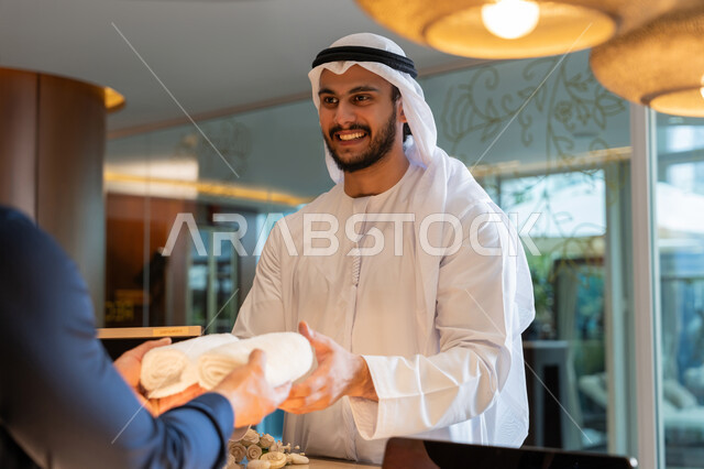 الاستمتاع بالعطلة الصيفية, الفنادق والمنتجعات في الإمارات العربية المتحدة, قضاء الأوقات في الإجازة, رجل عربي خليجي إماراتي مبتسم يرتدي الكندورة والغترة البيضاء يقوم بأخذ المناشف من مكتب الاستقبال, رحلات الصيف الترفيهية