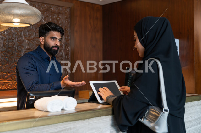 استخدام التقنيات الحديثة والمتطورة, القيام بالحجز الالكتروني, قضاء الأوقات الممتعة في العطلة الصيفية, امرأة عربية خليجية إماراتية محجبة ترتدي العباءة السوداء تقوم بإجراءات الحجز والدخول لدى مكتب الاستقبال في الفندق عن طريق الجهاز اللوحي