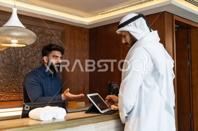 القيام بالحجز الالكتروني, استخدام التقنيات الحديثة والمتطورة, قضاء الأوقات الممتعة في العطلة الصيفية, رجل عربي خليجي إماراتي يرتدي الكندورة والغترة البيضاء يقوم بإجراءات الحجز والدخول لدى مكتب الاستقبال في الفندق عن طريق الجهاز اللوحي