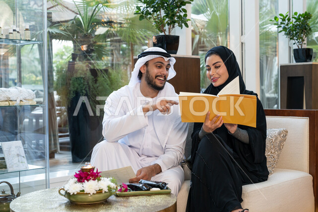 الإشارة بإصبع السبابة, قضاء الأوقات الممتعة في العطلة الصيفية, الأجواء العائلية السعيدة, القيام بالأنشطة الترفيهية في الإجازة, رجل عربي خليجي إماراتي يرتدي الكندورة والغترة البيضاء يجلس مع زوجته في إحدى المنتجعات ويقومان بالاختيار من القائمة