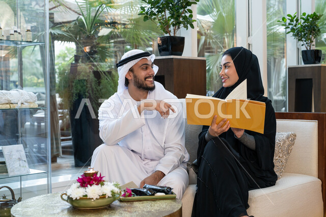Having fun in the summer vacation, happy family atmosphere, doing recreational activities on vacation, an Emirati Gulf Arab man wearing a white kandura and ghutra sitting with his wife in a resort and choosing from the menu