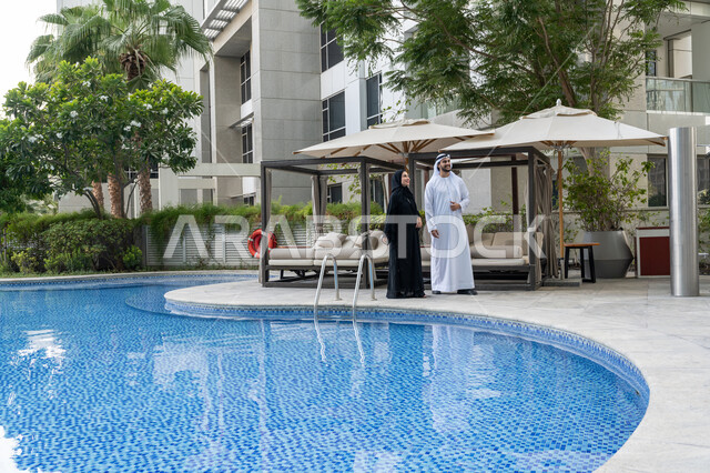 الاستمتاع بالعطلة الصيفية على المسبح, قضاء الأوقات الممتعة مع العائلة, الأجواء العائلية السعيدة, القيام بالأنشطة الترفيهية في الإجازة, رجل عربي خليجي إماراتي يرتدي الكندورة والغترة البيضاء يقف مع زوجته ويستمتعان في إحدى المنتجعات في الإمارات