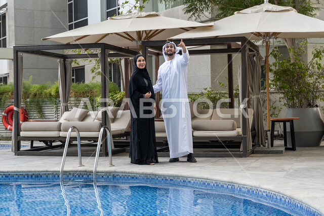 الاستمتاع بالعطلة الصيفية على المسبح, قضاء الأوقات الممتعة مع العائلة, الأجواء العائلية السعيدة, القيام بالأنشطة الترفيهية في الإجازة, رجل عربي خليجي إماراتي يرتدي الكندورة والغترة البيضاء يقف مع زوجته ويستمتعان في إحدى المنتجعات في الإمارات