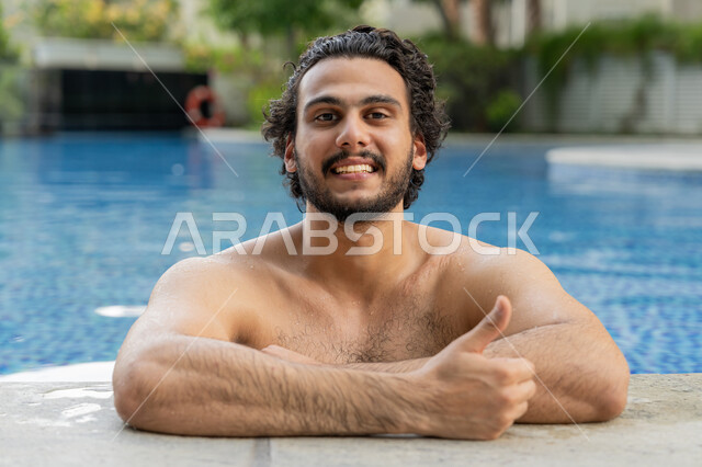 تعابير الجودة والامتياز, ممارسة رياضة السباحة الممتعة, القيام بالأنشطة الترفيهية المسلية, مفهوم الاستجمام والاختلاء, الجلوس والاسترخاء في المسبح, صورة مقربة لشاب عربي خليجي إماراتي يجلس داخل حوض السباحة يشير بإصبع لإبهام للأعلى