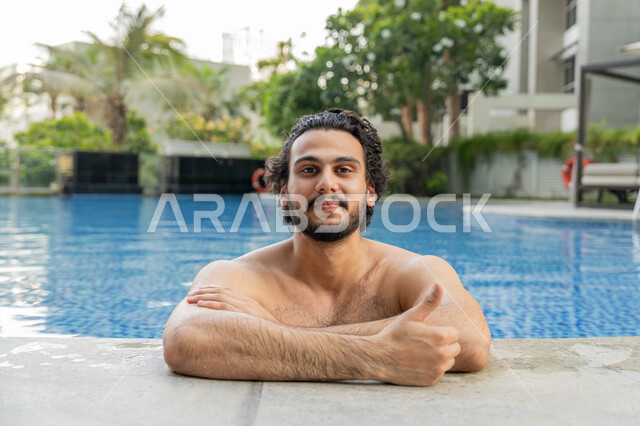 تعابير الجودة والامتياز, ممارسة رياضة السباحة الممتعة, القيام بالأنشطة الترفيهية المسلية, مفهوم الاستجمام والاختلاء, الجلوس والاسترخاء في المسبح, صورة مقربة لشاب عربي خليجي إماراتي يجلس داخل حوض السباحة يشير بإصبع لإبهام للأعلى