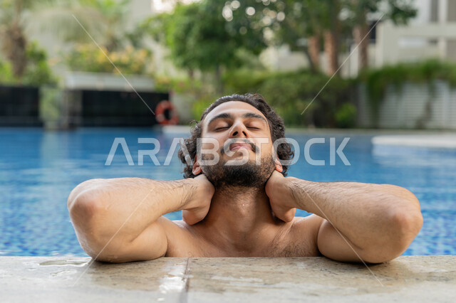 القيام بالأنشطة الترفيهية المسلية, مفهوم الاستجمام والاختلاء, ممارسة رياضة السباحة الممتعة, الجلوس والاسترخاء في المسبح, صورة مقربة لشاب عربي خليجي إماراتي يجلس داخل حوض السباحة في إحدى المنتجعات بالإمارات