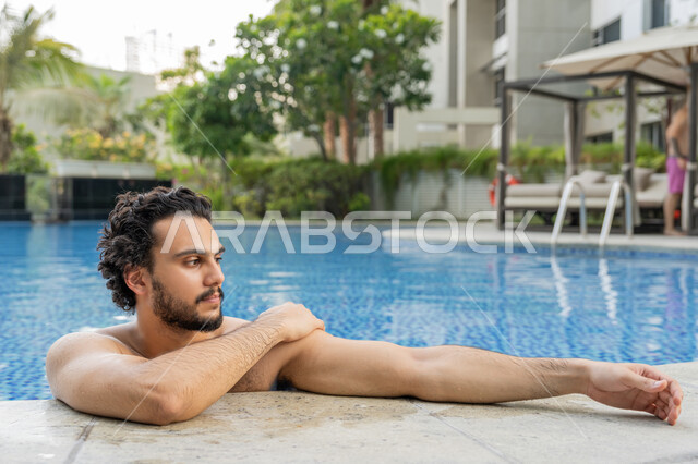 ممارسة رياضة السباحة الممتعة, الجلوس والاسترخاء في المسبح, القيام بالأنشطة الترفيهية المسلية, مفهوم الاستجمام والاختلاء, صورة مقربة لشاب عربي خليجي إماراتي يجلس داخل حوض السباحة في إحدى المنتجعات بالإمارات