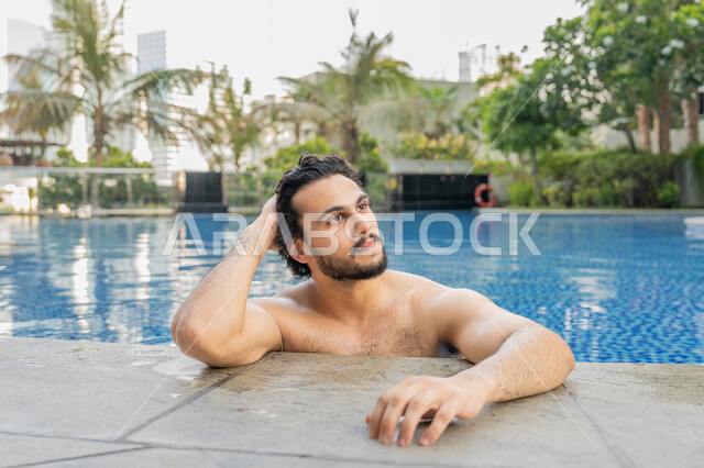 القيام بالأنشطة الترفيهية المسلية, مفهوم الاستجمام والاختلاء, ممارسة رياضة السباحة الممتعة, الجلوس والاسترخاء في المسبح, صورة مقربة لشاب عربي خليجي إماراتي يجلس داخل حوض السباحة في إحدى المنتجعات بالإمارات