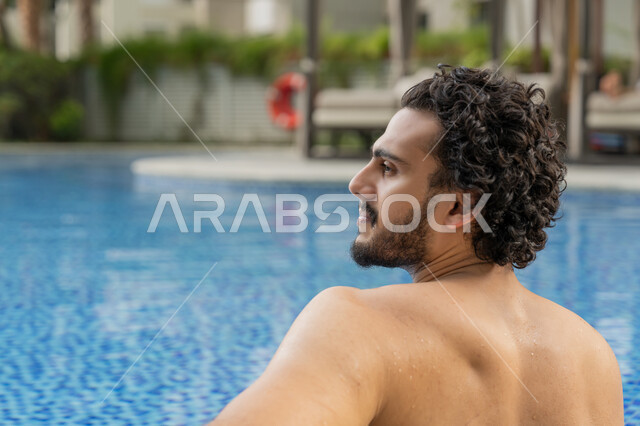 مفهوم الاستجمام والاختلاء, الجلوس والاسترخاء في المسبح, ممارسة رياضة السباحة الممتعة, القيام بالأنشطة الترفيهية المسلية, صورة مقربة من الخلف لشاب عربي خليجي إماراتي يجلس داخل حوض السباحة في إحدى المنتجعات بالإمارات