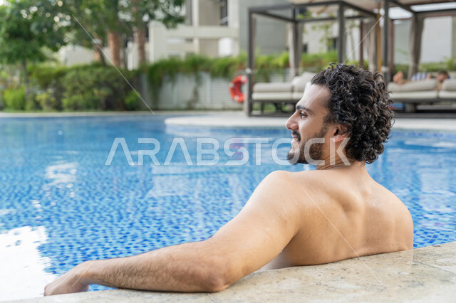 الجلوس والاسترخاء في المسبح, مفهوم الاستجمام والاختلاء, ممارسة رياضة السباحة الممتعة, القيام بالأنشطة الترفيهية المسلية, صورة مقربة من الخلف لشاب عربي خليجي إماراتي يجلس داخل حوض السباحة في إحدى المنتجعات بالإمارات