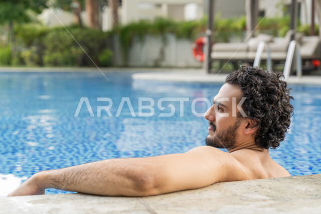 مفهوم الاستجمام والاختلاء, الجلوس والاسترخاء في المسبح, ممارسة رياضة السباحة الممتعة, القيام بالأنشطة الترفيهية المسلية, صورة مقربة من الخلف لشاب عربي خليجي إماراتي يجلس داخل حوض السباحة في إحدى المنتجعات بالإمارات