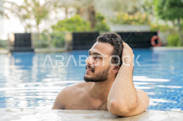 الجلوس والاسترخاء في المسبح, ممارسة رياضة السباحة الممتعة, القيام بالأنشطة الترفيهية المسلية, مفهوم الاستجمام والاختلاء, صورة مقربة لشاب عربي خليجي إماراتي يجلس داخل حوض السباحة في إحدى المنتجعات بالإمارات