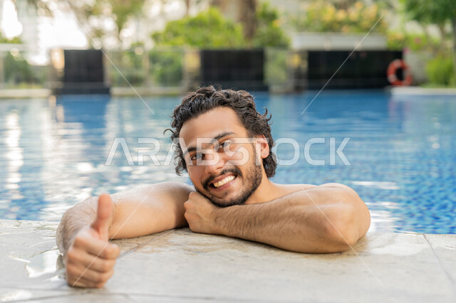النظر إلى الكاميرا بإيماءات السعادة والسرور, ممارسة رياضة السباحة الممتعة, القيام بالأنشطة الترفيهية المسلية, مفهوم الاستجمام والاختلاء, صورة مقربة لشاب عربي خليجي إماراتي يجلس داخل حوض السباحة يشير بإصبع لإبهام للأعلى بتعابير الجودة والامتياز