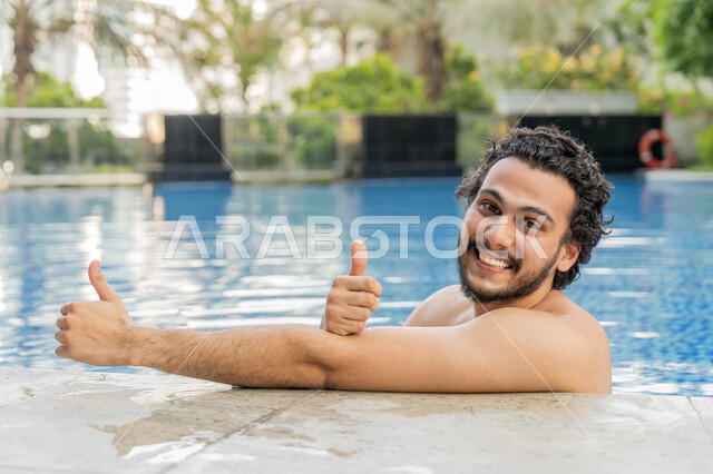 النظر إلى الكاميرا بإيماءات السعادة والسرور, ممارسة رياضة السباحة الممتعة, القيام بالأنشطة الترفيهية المسلية, مفهوم الاستجمام والاختلاء, صورة مقربة لشاب عربي خليجي إماراتي يجلس داخل حوض السباحة يشير بإصبع لإبهام للأعلى بتعابير الجودة والامتياز