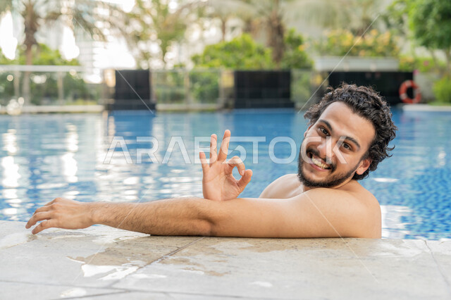 تعابير الجودة والامتياز, ممارسة رياضة السباحة الممتعة, القيام بالأنشطة الترفيهية المسلية, مفهوم الاستجمام والاختلاء, الجلوس والاسترخاء في المسبح, صورة مقربة لشاب عربي خليجي إماراتي يجلس داخل حوض السباحة وينظر إلى الكاميرا بإيماءات السعادة والسرور
