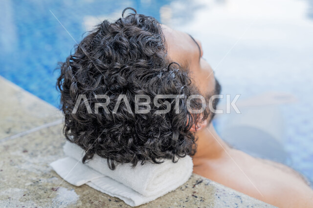 ممارسة رياضة السباحة الممتعة, القيام بالأنشطة الترفيهية المسلية, مفهوم الاستجمام والاختلاء, الجلوس والاسترخاء في المسبح, صورة مقربة لشاب عربي خليجي إماراتي يجلس داخل حوض السباحة في إحدى المنتجعات بالإمارات