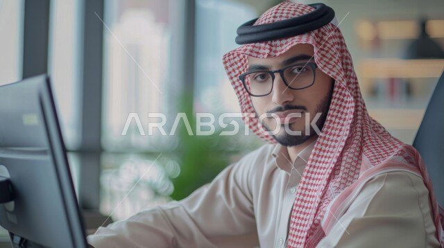 إدارة شؤون الأعمال وتنظيمها، استخدام الأجهزة التكنولوجية الحديثة والمتطورة، مهن ووظائف ادارية مكتبية, صورة مقربة من الجانب لموظف عربي خليجي سعودي يرتدي الثوب التقليدي والشماغ يقوم بالعمل على الحاسوب في داخل المكتب