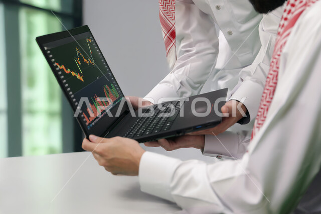 إنجاز الأعمال المكتبية، التفاهم والتعاون بين الزملاء لتحقيق أهداف الشركة، التحقق والتدقيق في البيانات المالية واسهم البورصة، تطوير خطة وآلية العمل، صورة مقربة لرجل عربي خليجي سعودي يرتدي الزي التقليدي يقوم بشرح المهام لزميله عبر جهاز الحاسوب المحمول