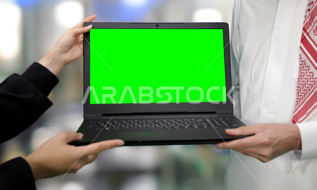 إنجاز مهام العمل عبر الإنترنت بين زملاء العمل، القيام بالدردشات والمحادثات مع العائلة والأصدقاء، تصوير مقرب ليدين رجل وامرأة عربيان خليجيان سعوديان يحملان جهاز الحاسوب المحمول، أجهزة إلكترونية بشاشة خضراء فارغة, كروما