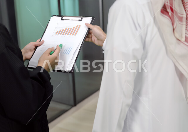 Writing and recording notes, completing office work, understanding and cooperation between colleagues to achieve company goals, verifying and auditing financial offers, developing a work plan and mechanism, close-up of a Saudi Arabian Gulf woman wearing a black abaya explaining tasks to her colleague