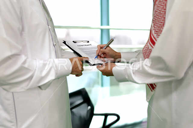 كتابة وتسجيل الملاحظات, إنجاز الأعمال المكتبية، التفاهم والتعاون بين الزملاء لتحقيق أهداف الشركة، التحقق والتدقيق في العروض المالية، تطوير خطة وآلية العمل، صورة مقربة لرجل عربي خليجي سعودي يرتدي الزي التقليدي يقوم بشرح المهام لزميله