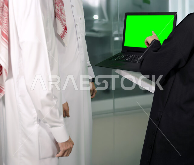ادارة اعمال الشركة بين زملاء العمل, عرض شاشة خضراء فارغه عبر الاجهزة المتطورة، صورة مقربة لامراة عربية خليجية سعودية ترتدي العباءة السوداء تشير باصبع السبابة الى جهاز الحاسوب المحمول، استخدام اللابتوب في الشركات, كروما