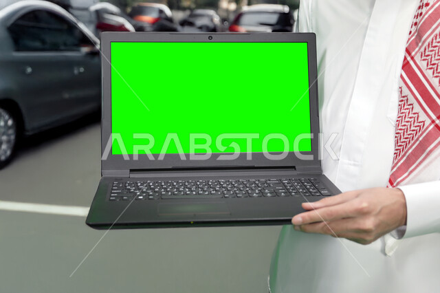 عرض شاشة خضراء فارغه عبر الاجهزة المتطورة، صورة مقربة لرجل عربي خليجي سعودي يرتدي الثوب التقليدي والشماغ يحمل بيده جهاز الحاسوب المحمول، استخدام اللابتوب في تنظيم خطط العمل, كروما
