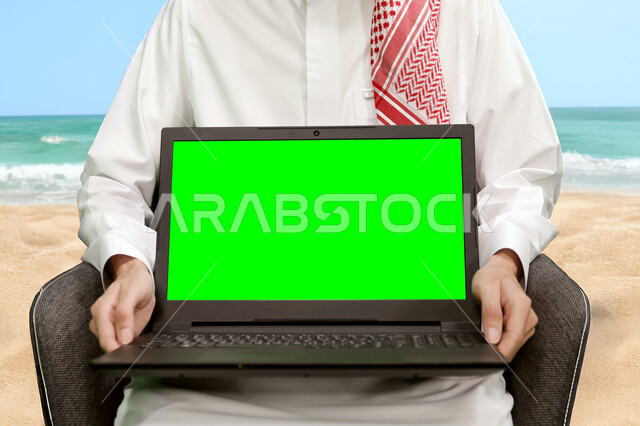 عرض شاشة خضراء فارغه عبر الاجهزة المتطورة، صورة مقربة لرجل عربي خليجي سعودي يرتدي الثوب التقليدي يجلس على الكرسي على شاطئ البحر يحمل بيده الحاسب المحمول، استخدام اللابتوب في تنظيم خطط العمل, كروما