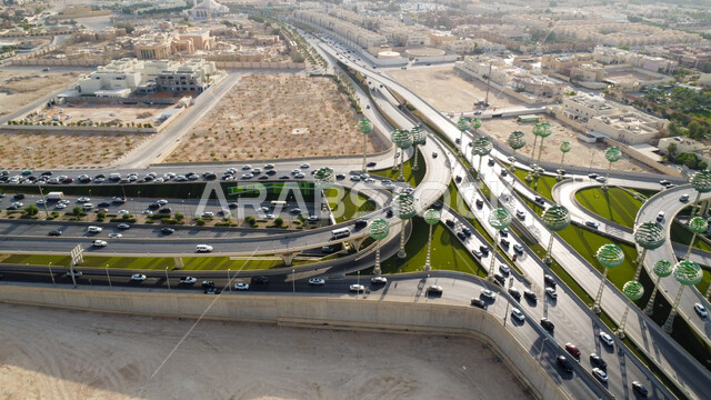 حركة سير السيارات على الطريق السريع في مدينة الرياض، الفن الهندسي المعماري للمجسمات الفنية على شكل أشجار خضراء، الاهتمام بزراعة النباتات الخضراء، الطرق المتقاطعة في دوار عرقة بالمملكة العربية السعودية، البنية التحتية المتطورة