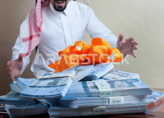 الأرباح العالية المجنية من إدارة الأعمال التجارة والبيع والشراء، المنافسة في سوق البورصة والاستثمارات المالية، رجل عربي خليجي سعودي يرتدي الثوب التقليدي والشماغ يقف بفرح أمام كومة من الأوراق المالية الموضوعة فوق بعضها، استغلال الفرص وعقد الصفقات الرابحة