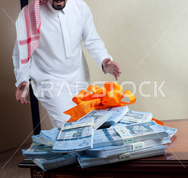 الأرباح العالية المجنية من إدارة الأعمال التجارة والبيع والشراء، المنافسة في سوق البورصة والاستثمارات المالية، رجل عربي خليجي سعودي يرتدي الثوب التقليدي والشماغ يقف بفرح أمام كومة من الأوراق المالية الموضوعة فوق بعضها، استغلال الفرص وعقد الصفقات الرابحة