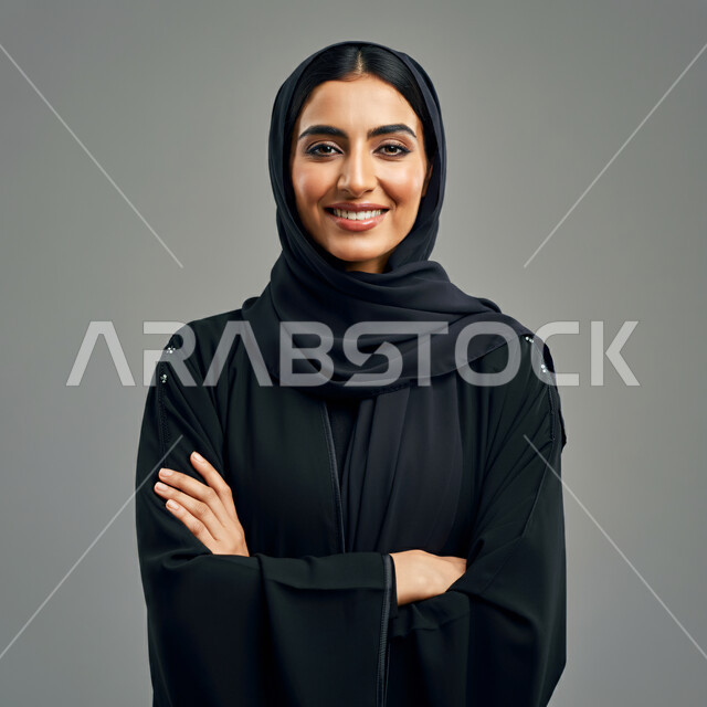  أزياء محجبات عصرية راقية، الملامح العربية الأصيلة، مفهوم الثقة بالنفس والاهتمام بالمظهر الخارجي، بورتريه مقرب لامرأة عربية خليجية سعودية مبتسمة ترتدي الحجاب والعباءة تنظر الى الكاميرا بتعابير السرور تقف بأيدي متقاطعة، خلفية رمادية