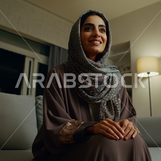  الهدوء والاسترخاء والراحة، أزياء محجبات عصرية راقية، مفهوم الأناقة والاهتمام بالمظهر الخارجي، صورة مقربة لامرأة عربية خليجية سعودية مبتسمة ترتدي العباءة والحجاب تنظر الى شيء ما بإيماءات السرور و الثقة بالنفس، قضاء اوقات ممتعة في المنزل
