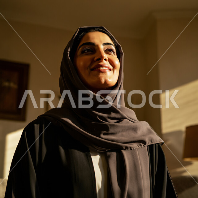 قضاء اوقات ممتعة في المنزل، الهدوء والاسترخاء والراحة، أزياء محجبات عصرية راقية، مفهوم الأناقة والاهتمام بالمظهر الخارجي، صورة مقربة لامرأة عربية خليجية سعودية مبتسمة ترتدي العباءة والحجاب تنظر الى شيء ما بإيماءات السرور و الثقة بالنفس