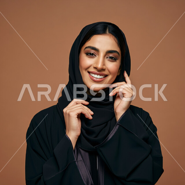 مفهوم النعومة والأنوثة، أزياء محجبات عصرية راقية، الأناقة والاهتمام بالمظهر الخارجي، بورتريه مقرب لامرأة عربية خليجية سعودية مبتسمة ترتدي العباءة والحجاب تنظر الى الكاميرا بإيماءات السرور و الثقة بالنفس، خلفية بنية 