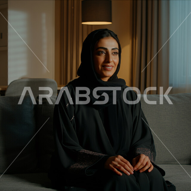 قضاء اوقات ممتعة في المنزل، الهدوء والاسترخاء والراحة، أزياء محجبات عصرية راقية، مفهوم الأناقة والاهتمام بالمظهر الخارجي، صورة مقربة لامرأة عربية خليجية سعودية مبتسمة ترتدي العباءة والحجاب تنظر الى شيء ما بإيماءات السرور و الثقة بالنفس